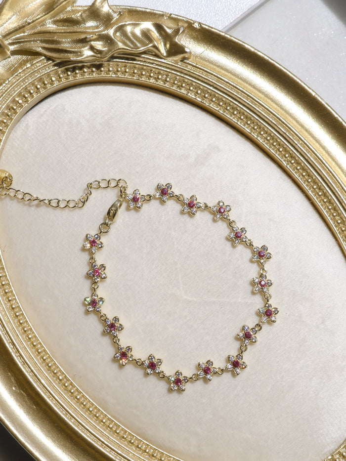 A gold chain bracelet with small flower motifs, each featuring a pink gemstone and clear rhinestones, displayed on a cream-colored surface inside an ornate gold frame.